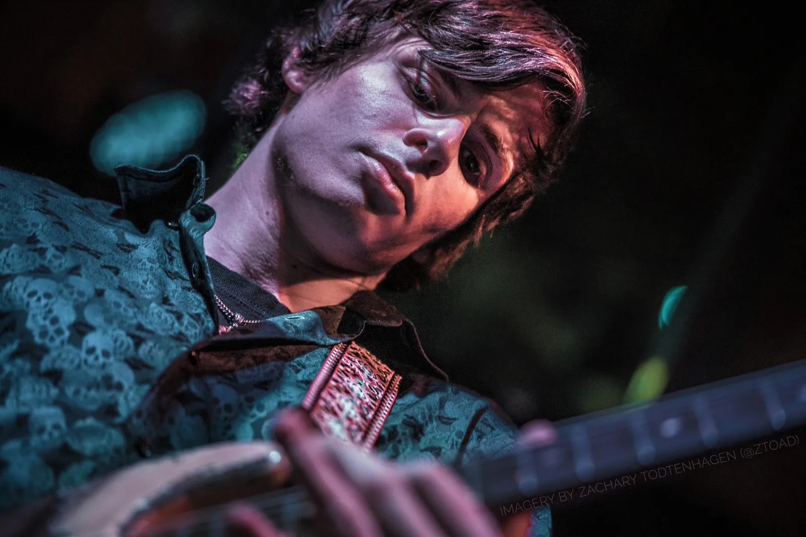 Close portrait of Hayden Fogle on stage, looking towards the camera over the neck of his guitar, lit in teal and warm pink stage light.