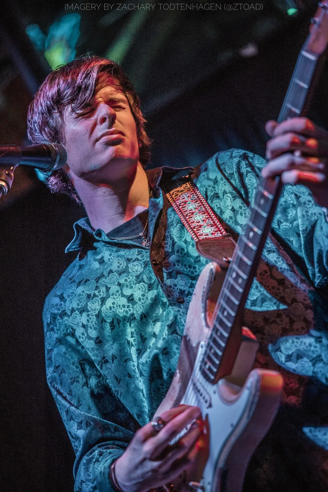 Hayden Fogle leaning into a microphone while playing, caught mid-phrase under magenta and teal stage lighting.