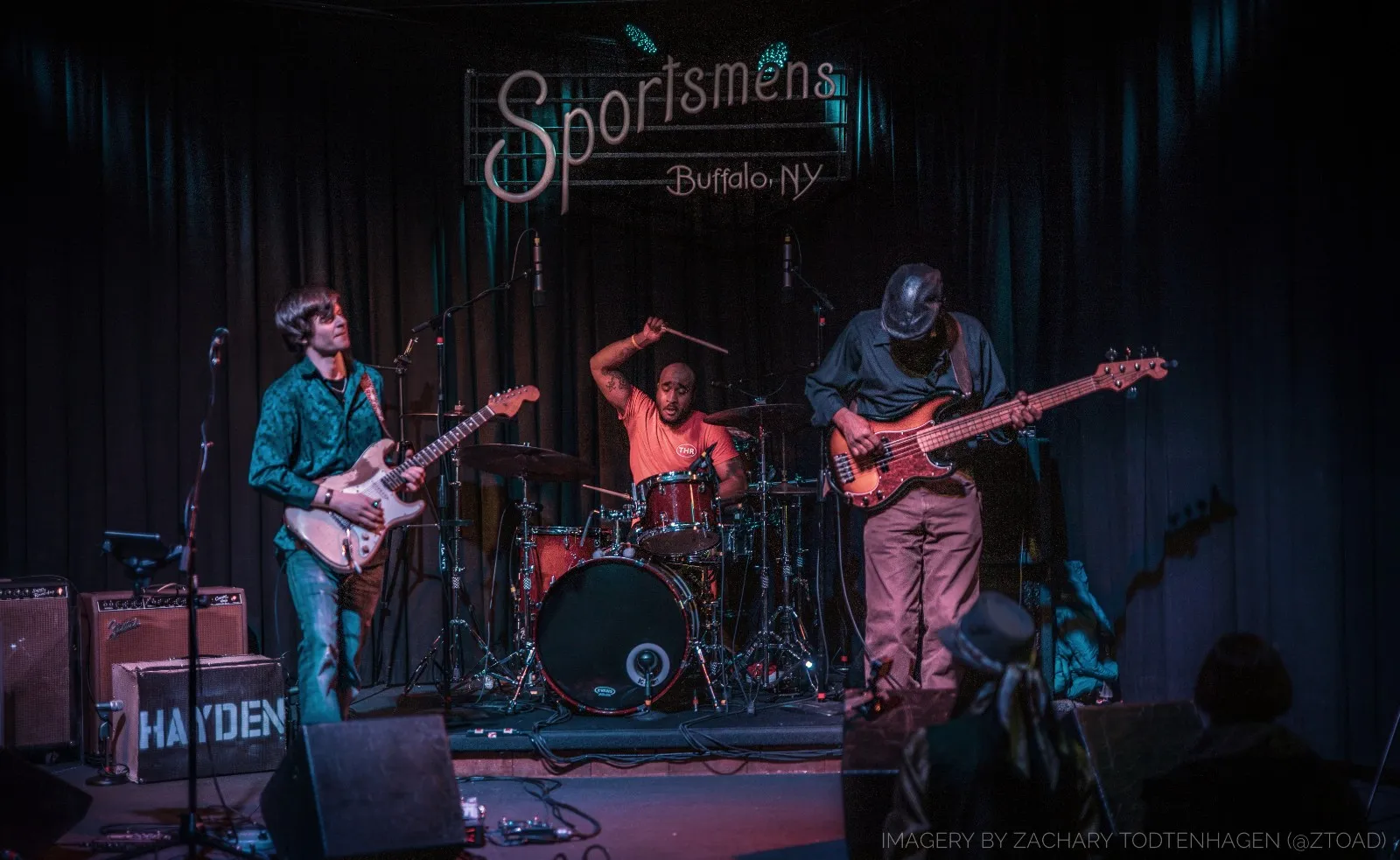 The Hayden Fogle Band as a trio on stage beneath the neon Sportsmens Buffalo NY sign, with guitar, drums and bass.