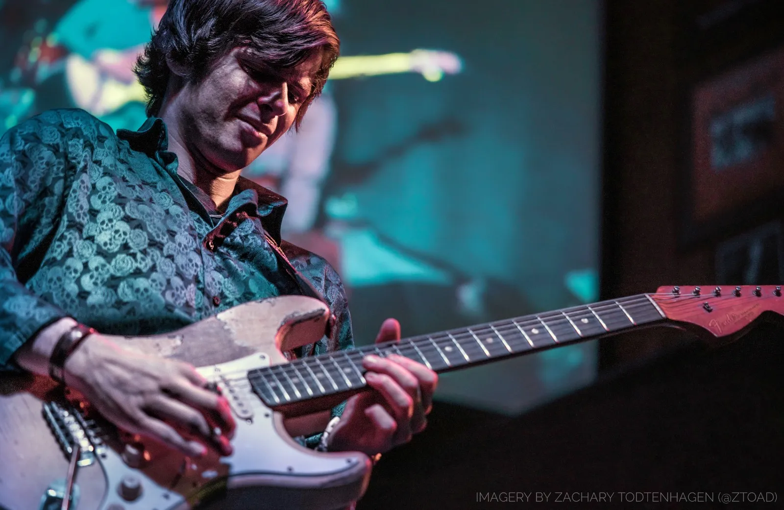 Hayden Fogle playing a worn Fender Stratocaster, eyes down on the fretboard, a projection screen glowing behind him.