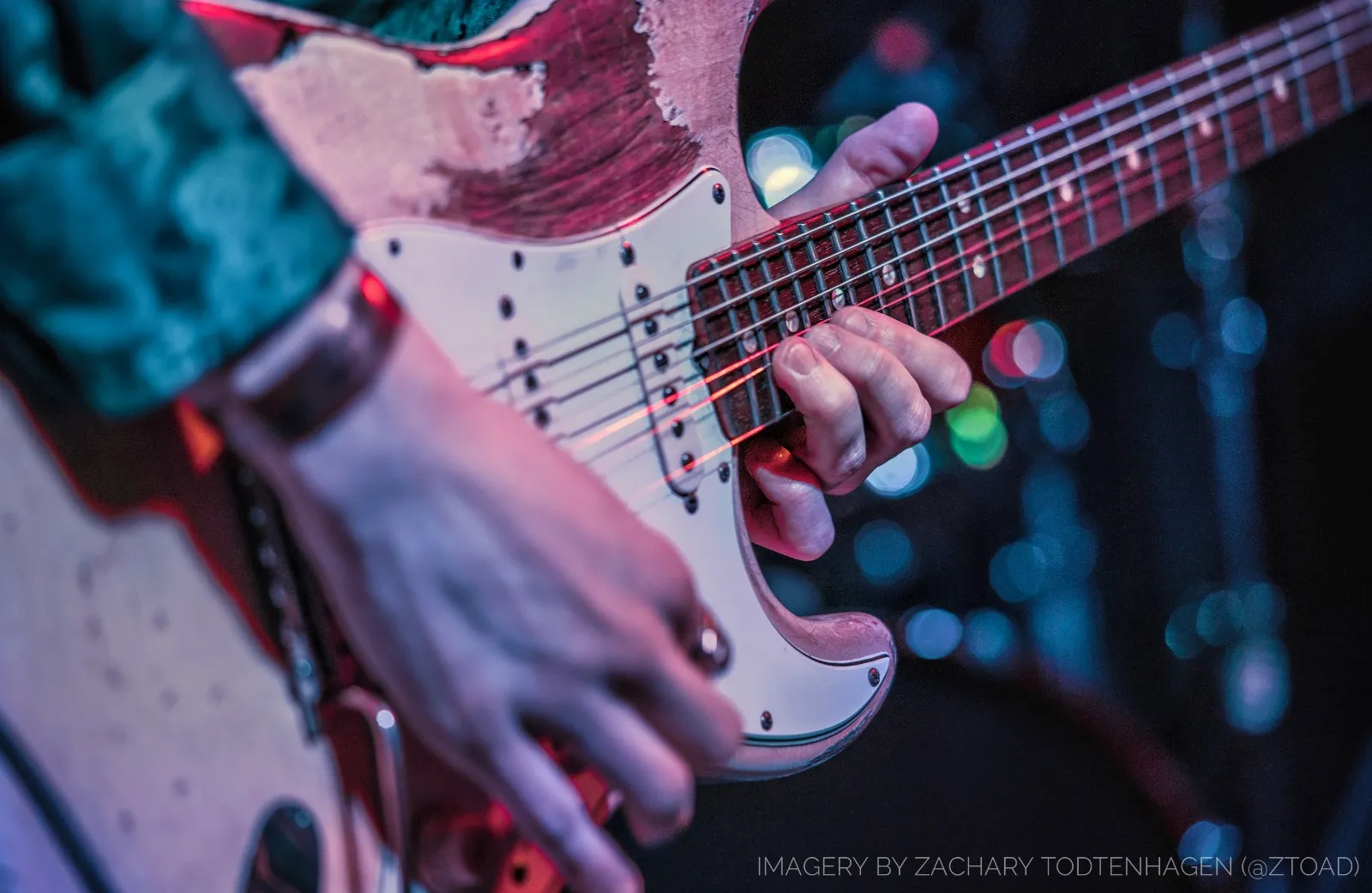 Detail of a battered sunburst Stratocaster in close-up, one hand fretting and one hand at the bridge, stage lights blurred behind.